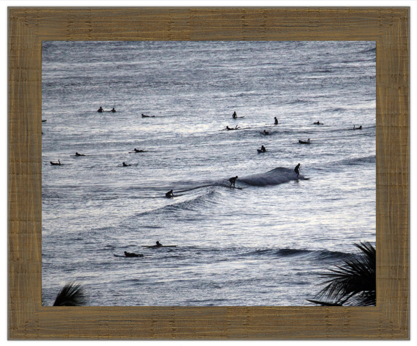 Framed artwork of ocean with surfers, framed in brown wood.