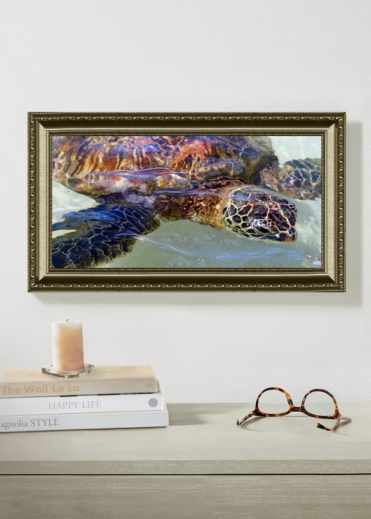 Framed painting of a turtle on a wall above books and glasses.