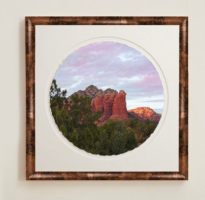 Framed circular artwork of a pink sunset, mountain landscape with a wooden frame.A collection of framed artwork prints with a deckled edge appearance, displayed in two museum style frames named Light Burlwood and Dark Burlwood. The frames are pictured in various sizes with a white mat and clear acrylic front.